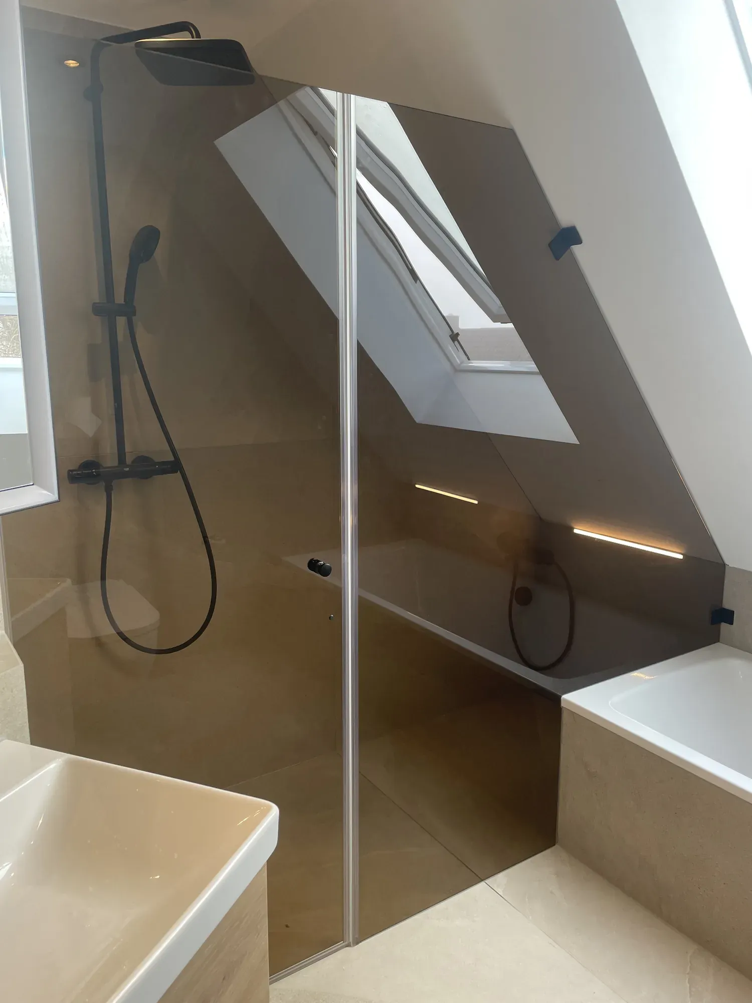 Neues Dachbad mit Glasdusche, sandfarbenen Fliesen und schwarzer Armatur unter dem Dachfenster