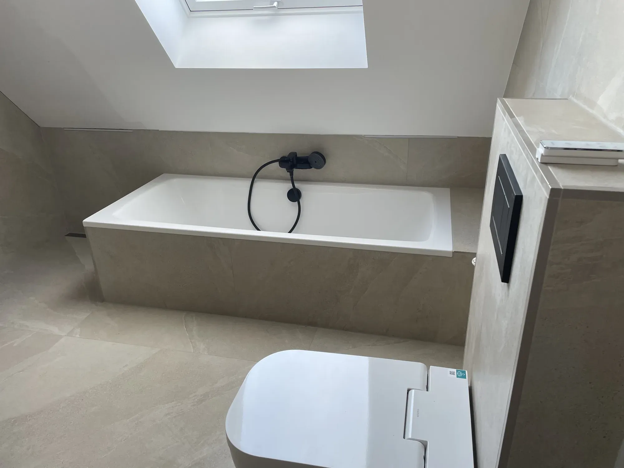 Badewanne unter der Dachschräge mit Dachfenster, sandfarbene Großformatfliesen und schwarze Armatur