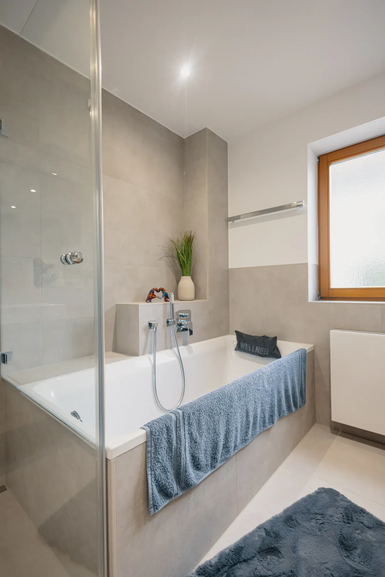 Badewanne mit Glasabtrennung, Handbrause und blauem Handtuch vor dem Fenster