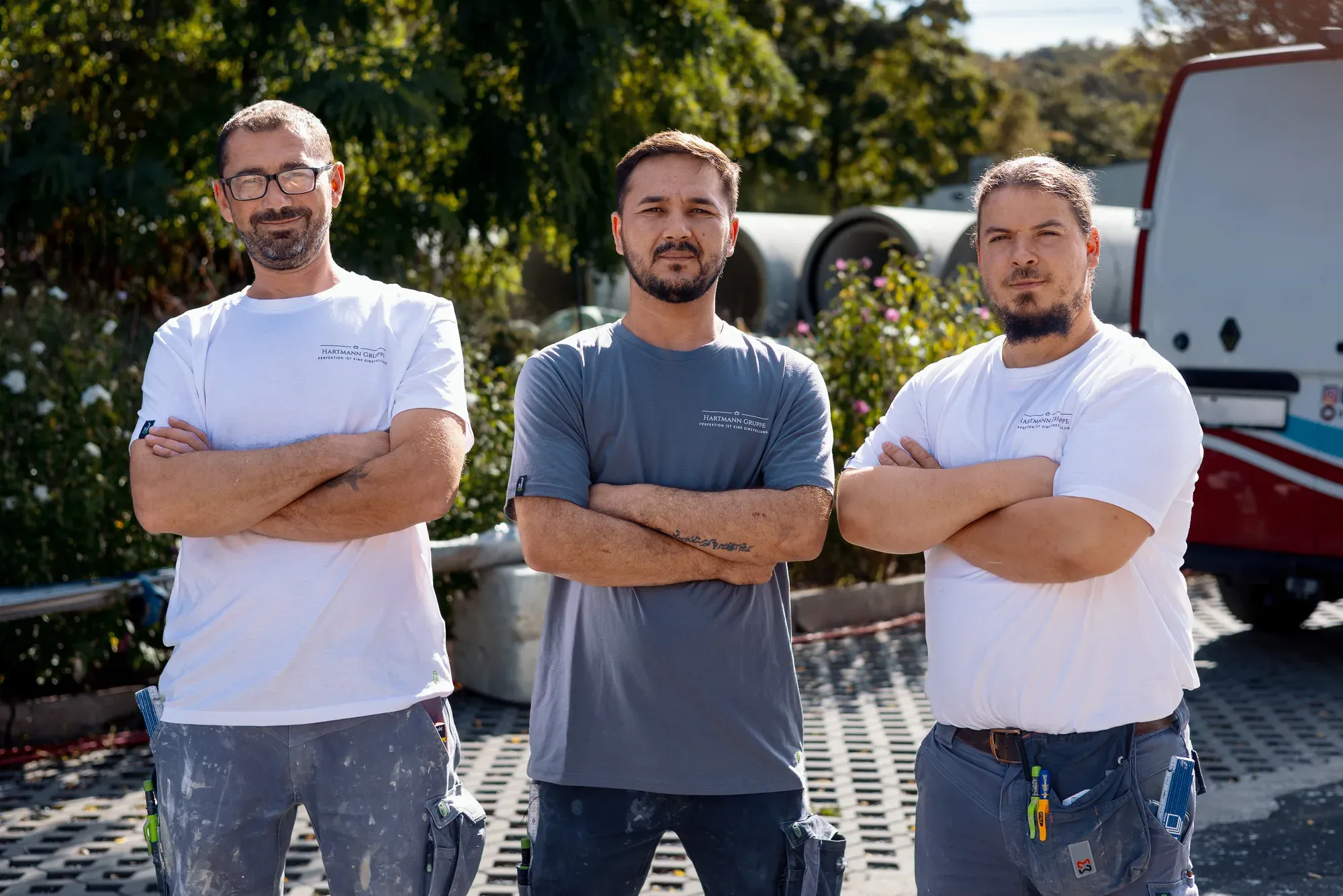 Drei Mitarbeiter der Hartmann Gruppe in Arbeitskleidung vor dem Firmenwagen
